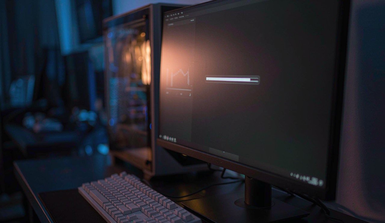 A gaming desk at night with a download progress graph on the monitor and a PC tower beside it