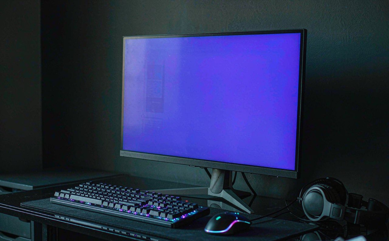 A dark gaming setup with a monitor glowing purple and an RGB keyboard, photographed at an angle