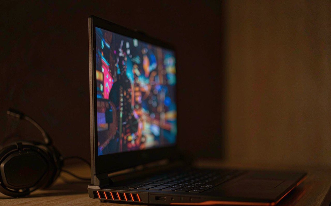 An open gaming laptop on a desk showing a colorful blurred game scene, with its glowing side vents visible in a dim room