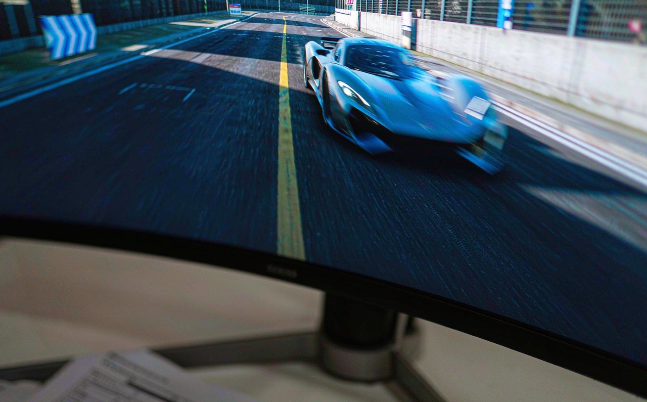 Close-up of a fast-moving esports racing game on a gaming monitor, with slight motion trail visible behind a car, next to the monitor spec sheet on paper